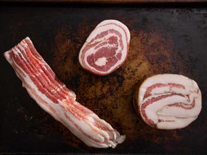 Overhead view of bacon, pancetta, and guanciale arranged on a well-seasoned baking sheet.