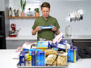 Sasha Marx from Serious Eats standing in a kitchen with a pile of different packages of pastas on the counter in front of him.