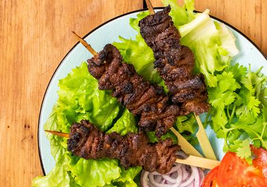 A plate with three Nigerian beef suya (spiced grilled skewers) on a bed of lettuce with sliced red onions, tomato sliced, and cilantro. 