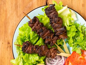 A plate with three Nigerian beef suya (spiced grilled skewers) on a bed of lettuce with sliced red onions, tomato sliced, and cilantro. 