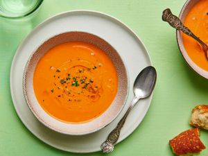 A bowl of Andalusian gazpacho in a ceramic bowl set on a plate. On the right hand periphery are another bowl of soup and a couple of pieces of torn bread.