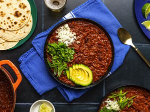 A large bowl of vegetarian bean chili topped with sliced avocado, scallions, cilantro, and white onion. The periphery of the image contains a wide variety of plates and bowls holding tortillas, sliced limes, and additional chili.