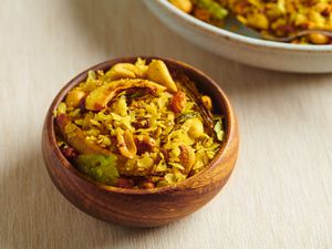 A small round wooden bowl holding poha chivda. There is a larger cream-colored ceramic bowl in the top right corner of the image.