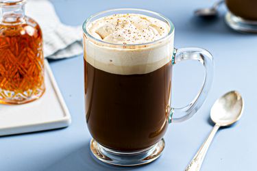 Spiced rum coffee with butterscotch whipped cream in a glass mug. There is a silver spoon to the right of the mug, and a small glass vessel of rum on the lefthand side of the image.