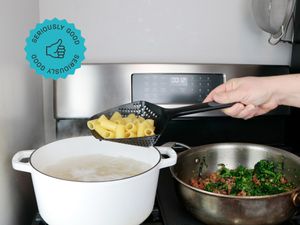 A colander spoon holding drained rigatoni of a Dutch oven full of boiling water