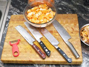 four nakiri knives on a cutting board with a bowl of cut up sweet potatoes