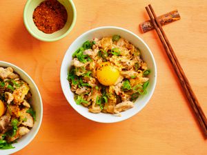 Oyakodon, in a white ceramic bowl alongside wooden chopsticks, a small ceramic bowl holding togarashi seasoning, and an additional bowl of oyakodon off to the left side.