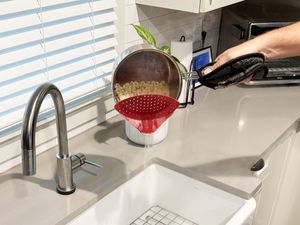 A person using a clip-on strainer to water from pasta noodles into a sink.