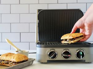 a person placing a sandwich onto a panini press and a small sheet tray holding another sandwich sitting besides the panini press