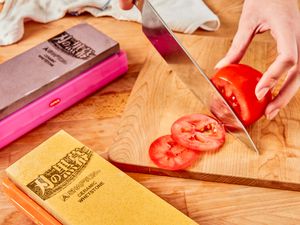 A person slices a tomato with a chef's knife. Two knife sharpeners are also in the frame.