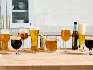 Numerous beer glasses on a kitchen countertop. Bottles and cans of beer are beside the glasses.