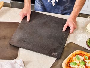 A person placing a pizza steel onto a kitchen countertop. A cooked pizza is on one of the pizza steels.