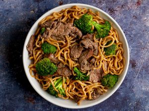 A white ceramic bowl holding lo mein with beef and broccoli on a purple colored stone surface.