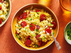 A bowl of fried rice with Chinese sausage. There is a silver spoon in the bowl. On the left periphery of the image is another bowl of fried rice. There is a glass of liquid in the top right corner, and a bowl holding sliced scallions on the right periphery of the image.