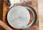 A metal tart pan on a wooden cutting board with pie dough and a rolling pin next to it