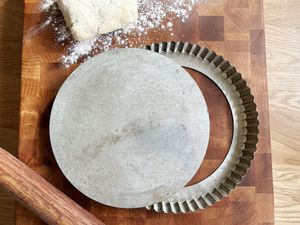 A metal tart pan on a wooden cutting board with pie dough and a rolling pin next to it