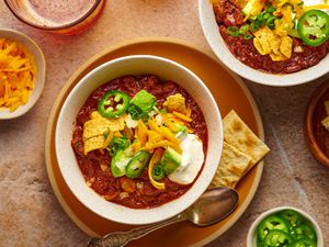 A white speckled ceramic bowl holding chili topped with corn chips, grated cheddar, sour cream, sliced jalapenos, avocado, and scallions. There are saltine crakers on the plate underneath the bowl of chili, and the periphery of the image has small bowls of additional toppings.