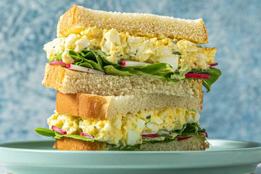 An egg salad sandwich, cut in half, with the two halves stacked on top of each other. The sandwich contains egg salad, butter lettuce, and thinly sliced radishes.