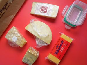 a variety of cheese on a red backdrop with parchment paper and a food container